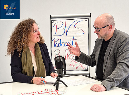 Newsbild Podcast Baurecht An einem weißen Tisch stehen sich 2 Personen im Profil gegenüber und diskutieren miteinander. Zwischen ihnen steht ein Mikrofon.