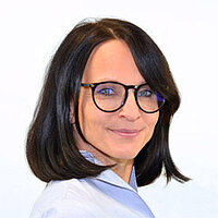 BVS Mitarbeiterin Sabrina Kirmayr Eine Frau mit schulterlangen schwarzen Haaren trägt eine hellblaue Bluse und eine dunkle Brille mit leicht ovalen Brillengläsern. Sie hat blaue Augen und lächelt mit geschlossenem Mund.