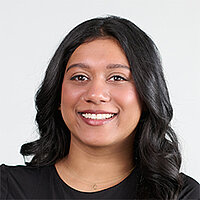 Man sieht das Gesicht einer jüngeren Frau mit braunen Augen und langen dunkelbraunen Haaren. Sie trägt ein braunes Sakko.