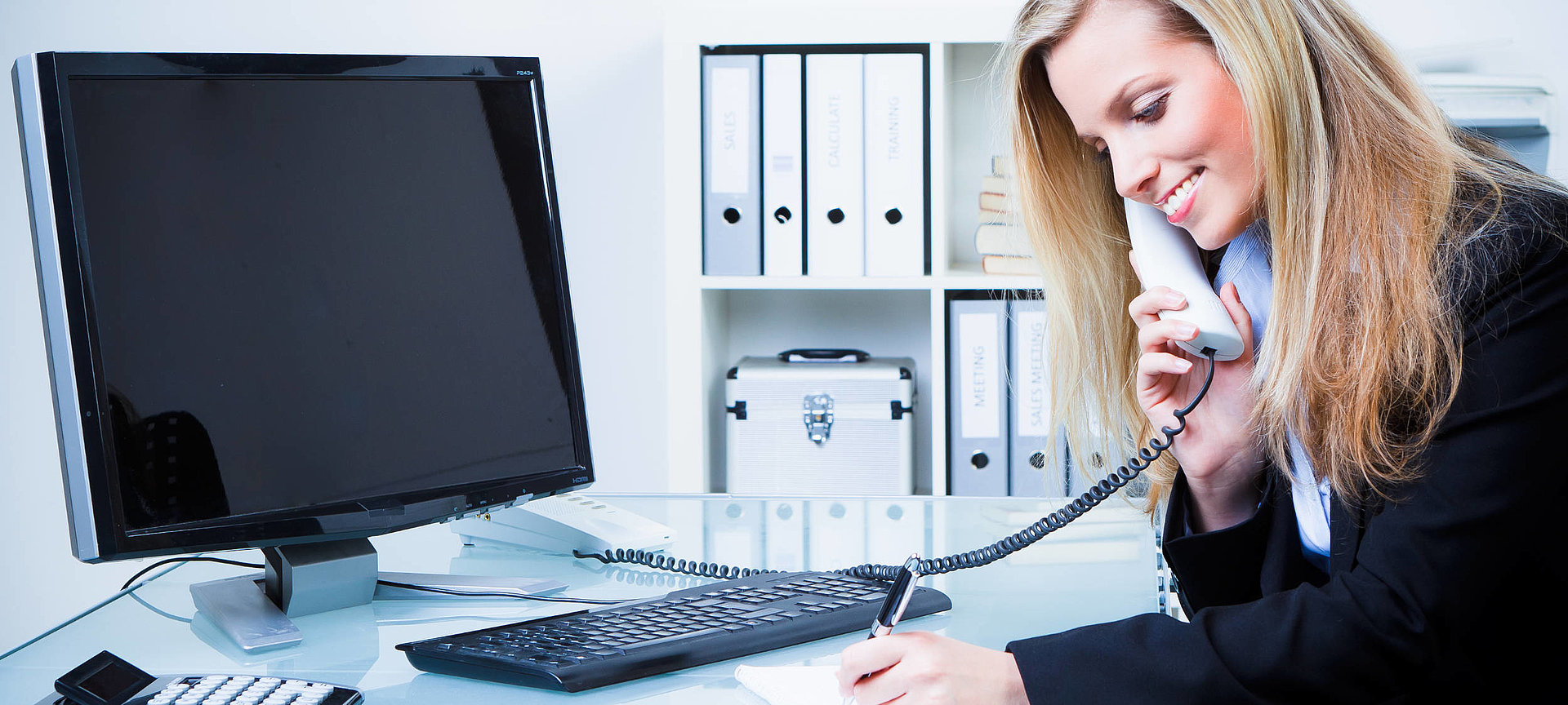 Das Bild ist das Übersichtsbild des Ausbildungsbereichs Verwaltung. Es zeigt eine junge Dame mit langen blonden Haaren, die an ihrem Schreibtisch sitzt und telefoniert. 