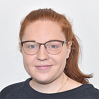 Maike Kaeßner Eine junge Frau mit Brille lund zurück gebundenen roten Haaren ist zu sehen. Sie trägt einen dunkelblauen Pullover.