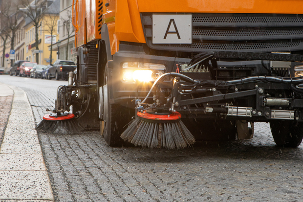 Themenbild Bauhofleitung Aus der Untersicht sieht man vom Straßenrand aus den linken unteren Teil einer Kehrmaschine mit beleuchteten Scheinwerfer.