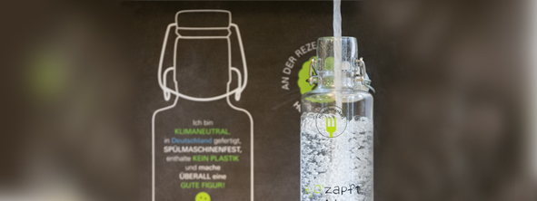 Themenbild Soulbottle "H2Ozapts is" Vor einem dunklen Hintergrund sieht man den oberen Teil einer gläsernen Trinkflasche mit Bügelverschluss in die sprudelndes Wasser eingefüllt wird.