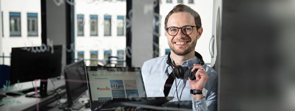 Themenbild Medienkompetenz Durch eine Glasscheibe sieht man angeschnitten den Oberkörper eines lächelnden Mannes, der um den Hals einen Kopfhörer trägt und in der Hand einen Laptop hält.