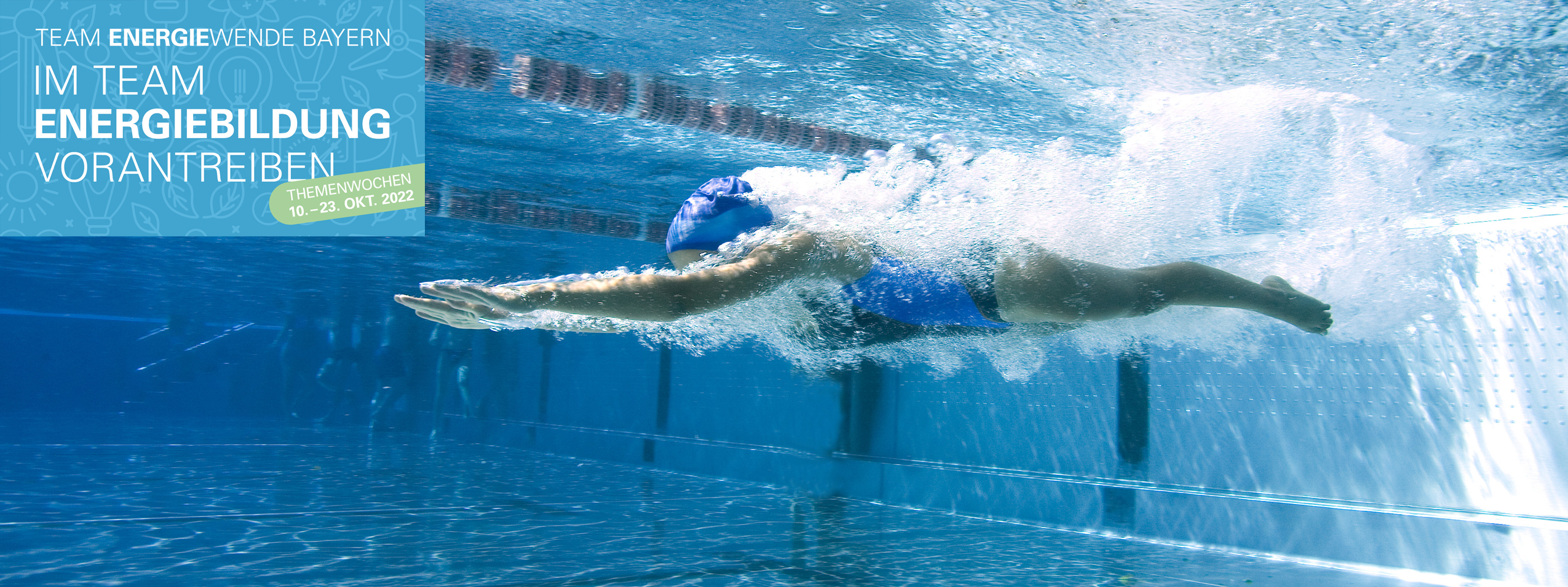 Eine Schwimmerin mit blauer Badehaube und blauem Badeanzug taucht unter Wasser in ein leeres Schwimmbecken.