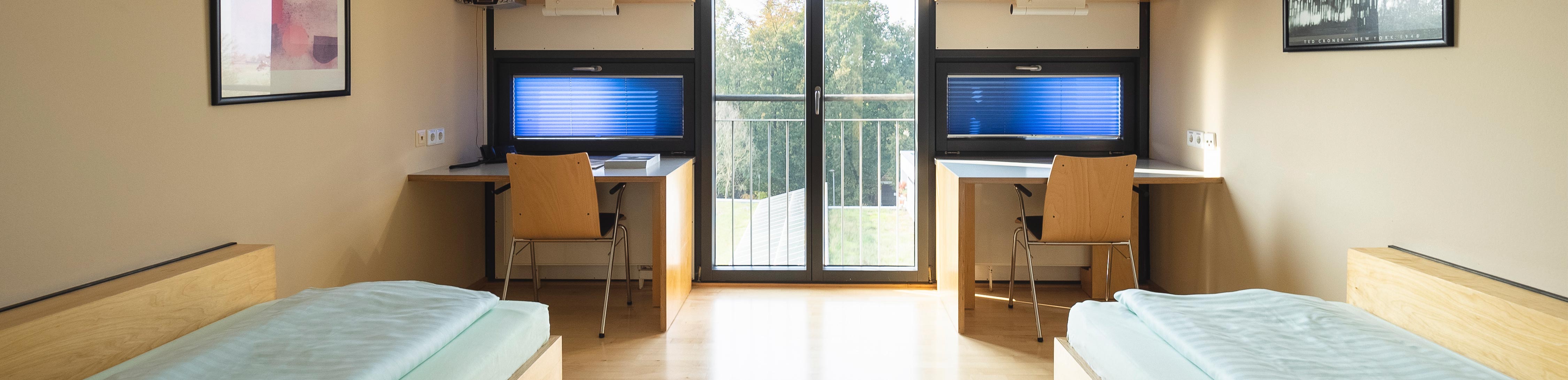 Das Bild ist das Headerbild der Zimmer im BVS-Bildungszentrum in Lauingen. Es zeigt ein Doppelzimmer von innen, das jeweils links und rechts spiegelverkehrt ein Bett und dahinter einen Tisch mit einem Stuhl stehen haben.