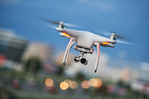 Eine weiße Drohne mit Kamera fliegt vor einer unscharfen Stadtsilhouette mit blauem Himmel.