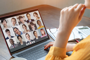 Am rechten Bildrand sieht man angeschnitten den Arm und das Kinn einer Frau in einer gelben Bluse. Sie sitzt an einem Tisch vor sich einen aufgeklappten Laptop. Auf dem Bildschirm sieht man viele kleine Fotos von Teilnehmenden in einem Onlinemeeting.