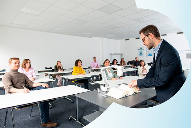 In einem Lehrsaal mit weißen Tischen sitzen Teilnehmende die auf einen Mann rechts schauen. Der Mann mit blauem Sakko und Brille blättert in einem Buch auf das eine Dokumentenkamera zeigt.
